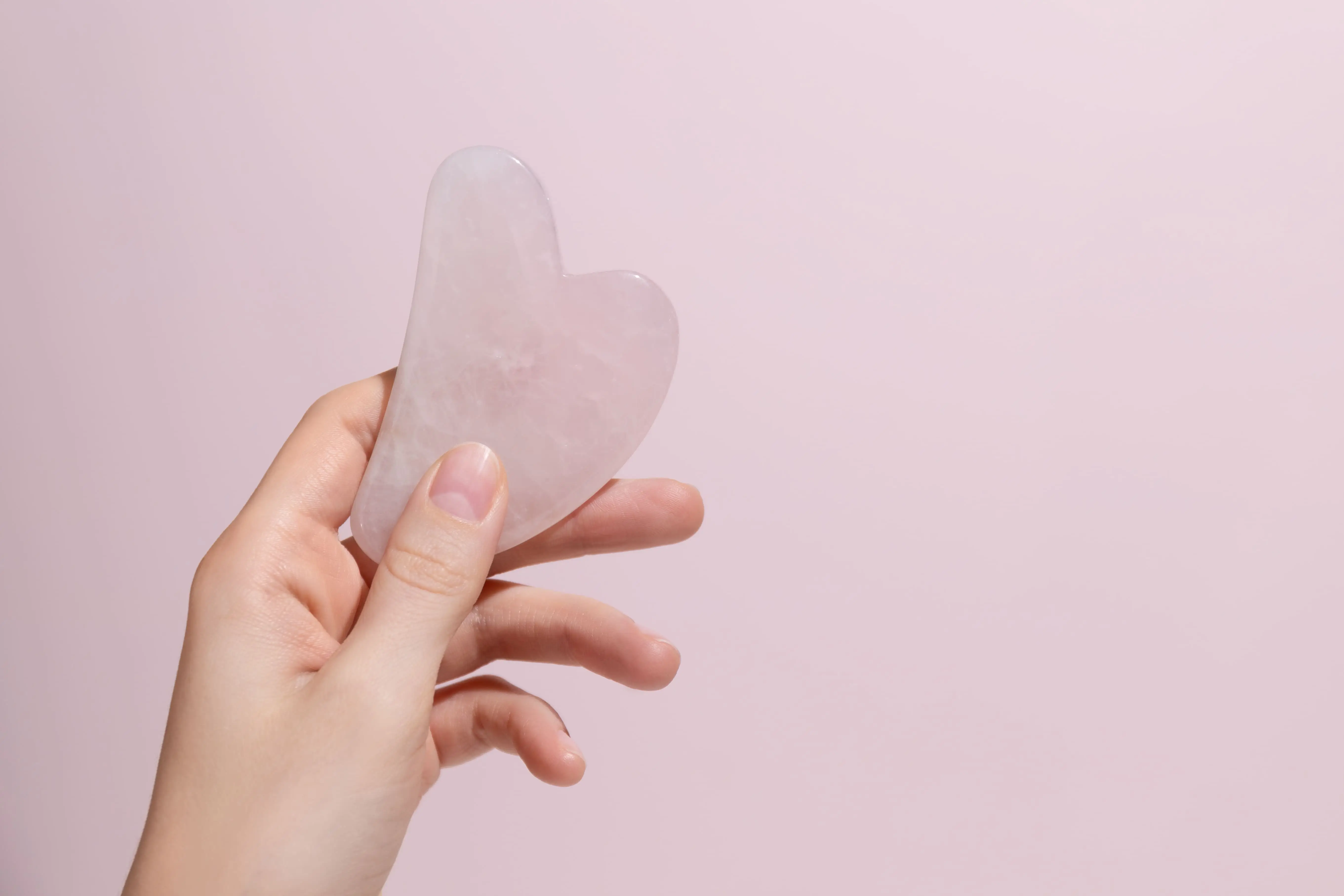 female-hand-holding-gua-sha-face-massager-pink-background-1